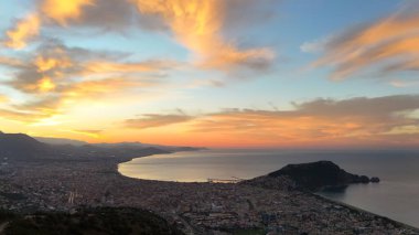 Türkiye 'nin Alanya kentinin geniş bir hava gündoğumu manzarası, şehri, körfezi ve kıyı şeridini sabahın erken saatlerinde sıcak ışıkla aydınlatıyor. Sakin deniz, uzak dağlar ve altın gökyüzü