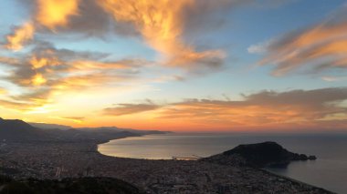 Türkiye 'nin Alanya kentinin geniş bir hava gündoğumu manzarası, şehri, körfezi ve kıyı şeridini sabahın erken saatlerinde sıcak ışıkla aydınlatıyor. Sakin deniz, uzak dağlar ve altın gökyüzü