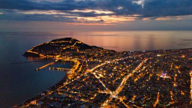 Türkiye 'nin Alanya kentinin insansız hava aracı tarafından ele geçirilmiş bir panoraması, aydınlık kenti, yarımadasını ve limanı sakin bir deniz ve alacakaranlık gökyüzüne karşı gösteriyor.