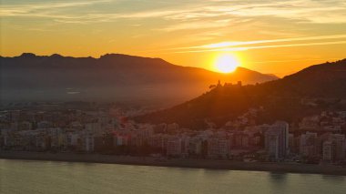 Günbatımında Cullera 'nın sinematik hava manzarası. Altın ışığa karşı kale ve kilise silueti var. Akdeniz kıyısı ve dağ manzarası dramatik bir kıyı kenti oluşturur..