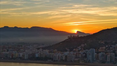 Günbatımında Cullera 'nın sinematik hava manzarası. Altın ışığa karşı kale ve kilise silueti var. Akdeniz kıyısı ve dağ manzarası dramatik bir kıyı kenti oluşturur..
