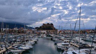 İspanya 'nın Akdeniz kıyısındaki Dnia limanı yelkenli ve yatlarla dolu. Tarihi kale tepesi, dramatik bulut oluşumları altında, rıhtımın üzerinde yükseliyor..