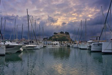 İspanya 'nın Akdeniz kıyısındaki Dnia limanı yelkenli ve yatlarla dolu. Tarihi kale tepesi, sakin suya yansıyan dramatik günbatımı bulutlarının altında, rıhtımın üzerinde yükseliyor..