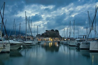 İspanya 'nın Akdeniz kıyısındaki Dnia limanı yelkenli ve yatlarla dolu. Tarihi kale tepesi, sakin suya yansıyan dramatik günbatımı bulutlarının altında, rıhtımın üzerinde yükseliyor..