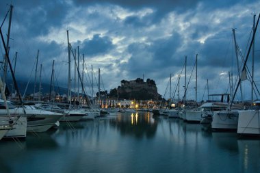 İspanya 'nın Akdeniz kıyısındaki Dnia limanı yelkenli ve yatlarla dolu. Tarihi kale tepesi, sakin suya yansıyan dramatik günbatımı bulutlarının altında, rıhtımın üzerinde yükseliyor..