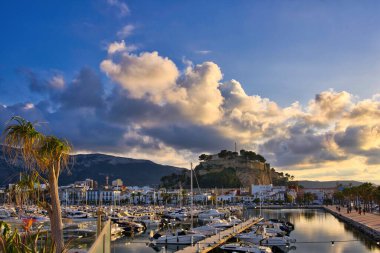 İspanya 'nın Akdeniz kıyısındaki Dnia limanında yelkenli tekneleri ve rıhtım güvertesiyle. Tarihi şato tepesi, sakin sulara yansıyan dramatik altın gün batımı bulutları altında limanın üzerinde yükseliyor..