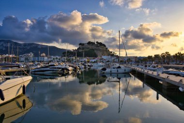 İspanya 'nın Akdeniz kıyısındaki Dnia limanında yelkenli tekneleri ve şehrin yukarısındaki tarihi şato tepesiyle. Altın günbatımı bulutları sakin liman suyuna yansıyarak sinematik bir sahil manzarası yaratıyor..