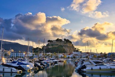 İspanya 'nın Akdeniz kıyısındaki Dnia limanında yelkenli tekneleri ve şehrin yukarısındaki tarihi şato tepesiyle. Altın günbatımı bulutları sakin liman suyuna yansıyarak sinematik bir sahil manzarası yaratıyor..