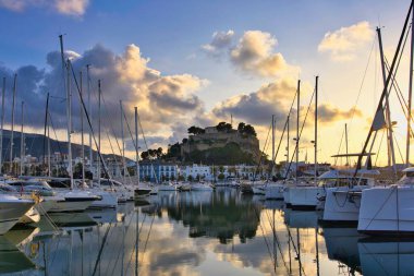 İspanya 'nın Akdeniz kıyısındaki Dnia limanında yelkenli tekneleri ve şehrin yukarısındaki tarihi şato tepesiyle. Altın günbatımı bulutları sakin liman suyuna yansıyarak sinematik bir sahil manzarası yaratıyor..