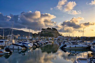 İspanya 'nın Akdeniz kıyısındaki Dnia limanında yelkenli tekneleri ve şehrin yukarısındaki tarihi şato tepesiyle. Altın günbatımı bulutları sakin liman suyuna yansıyarak sinematik bir sahil manzarası yaratıyor..