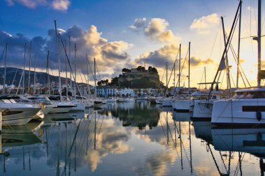 İspanya 'nın Akdeniz kıyısındaki Dnia limanında yelkenli tekneleri ve şehrin yukarısındaki tarihi şato tepesiyle. Altın günbatımı bulutları sakin liman suyuna yansıyarak sinematik bir sahil manzarası yaratıyor..
