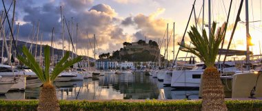 İspanya 'nın Akdeniz kıyısındaki Dnia limanında yelkenli tekneleri ve şehrin yukarısındaki tarihi şato tepesiyle. Altın günbatımı bulutları sakin liman suyuna yansıyarak sinematik bir sahil manzarası yaratıyor..