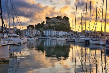 İspanya 'nın Akdeniz kıyısındaki Dnia limanında yelkenli tekneleri ve şehrin yukarısındaki tarihi şato tepesiyle. Altın günbatımı bulutları sakin liman suyuna yansıyarak sinematik bir sahil manzarası yaratıyor..