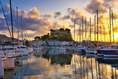 İspanya 'nın Akdeniz kıyısındaki Dnia limanında yelkenli tekneleri ve şehrin yukarısındaki tarihi şato tepesiyle. Altın günbatımı bulutları sakin liman suyuna yansıyarak sinematik bir sahil manzarası yaratıyor..