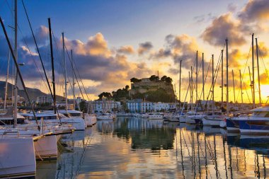 İspanya 'nın Akdeniz kıyısındaki Dnia limanında yelkenli tekneleri ve şehrin yukarısındaki tarihi şato tepesiyle. Altın günbatımı bulutları sakin liman suyuna yansıyarak sinematik bir sahil manzarası yaratıyor..