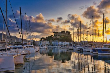 İspanya 'nın Akdeniz kıyısındaki Dnia limanında yelkenli tekneleri ve şehrin yukarısındaki tarihi şato tepesiyle. Altın günbatımı bulutları sakin liman suyuna yansıyarak sinematik bir sahil manzarası yaratıyor..