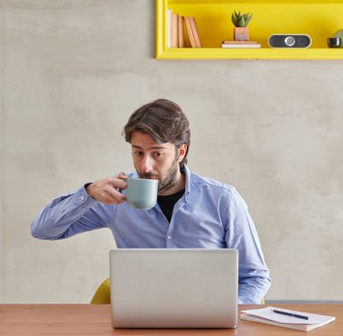 Adam dizüstü bilgisayarda, bir işte çalışıyor, kahve stili sarı kitaplık arka plan ve taş duvar içiyor..