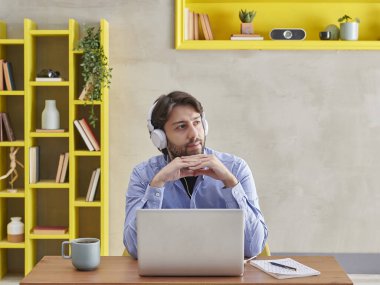 Adam videoda konuşuyor ya da dizüstü bilgisayarın önünde düşünüyor, gri taş duvar arkaplanı ve sarı kitaplık yuvası..