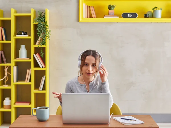 Woman is asking a question in video talking, grey wall background and yellow bookshelf, coffee laptop style.