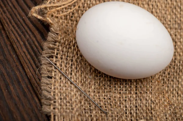 A chicken egg and a sewing needle on a homespun fabric with a rough texture. Close-up, selective focus.