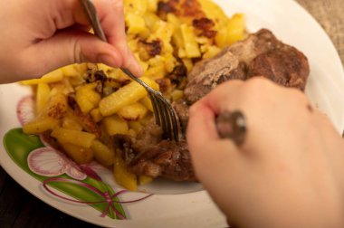 Birisi çatal ve bıçakla bir tabak kızarmış patatesle sulu bir parça et kesiyor. Seçici Odaklamayı Kapat