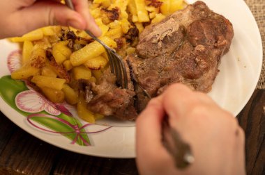 Birisi çatal ve bıçakla bir tabak kızarmış patatesle sulu bir parça et kesiyor. Seçici Odaklamayı Kapat