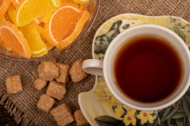 Cam bir vazoda çok renkli marmelat dilimleri, bir parça kahverengi kamış şekeri ve bir fincan çay, yakın plan, seçici bir odak noktası