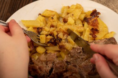 Birisi çatal ve bıçakla bir tabak kızarmış patatesle sulu bir parça et kesiyor. Seçici Odaklamayı Kapat