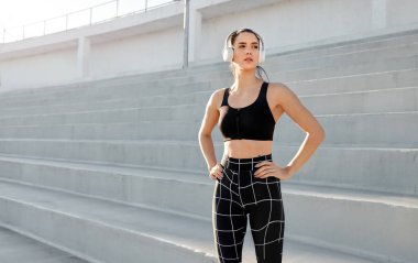 Spor kıyafetleri ve kulaklıklar takmış, elleri kalçasında spor salonunun merdivenlerinde duran genç ve zinde bir kadın.