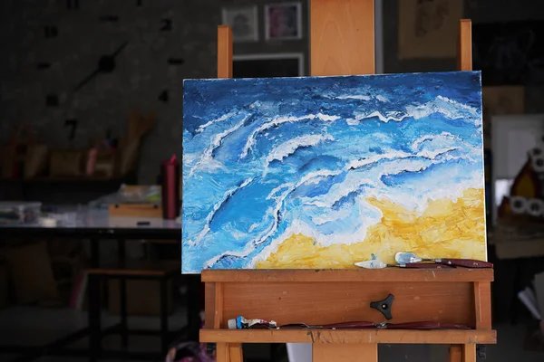 Acrylic sea and beach painting on drawing stand with palette and brush. Blue wave on golden sand hand painting with brush and knife palette technique. Art on canvas.