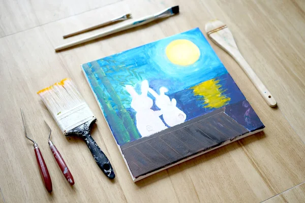 Acrylic art of two rabbit in love on roof under the moonlight at night, painting on drawing stand with brush. Blue wave on golden sand hand painting with brush technique. Art on canvas