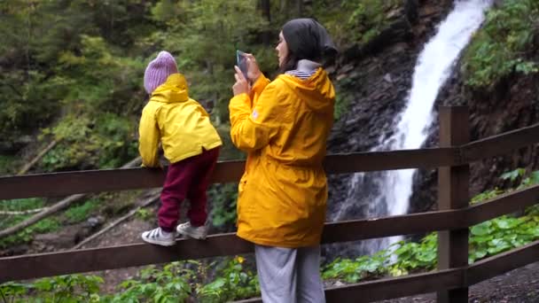 Anya gyermek öltözött sárga esőkabát áll a vízesés közelében — Stock videók