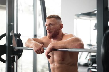 Spor salonundaki adam ağır antrenmandan sonra halterle dinleniyor. Beyaz tenli yakışıklı sporcu ara verdi. Spor aletleri, halter makinesinin yanında duran sportif adam. Mükemmel kaslı erkek vücudu ideal. Çıplak gövde.