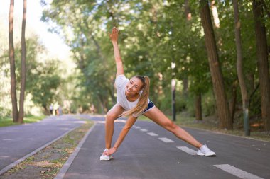Kadın koşucu, yaz parkında egzersiz yapmadan önce bacak esnetiyor. Orta yaşlı atletik kadın, koşuya çıkmadan önce vücudu ısıtıyor. Beyaz şortuyla koşuyor.