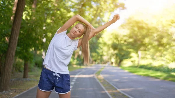 Woman runner stretching arms before exercising summer park  morning Middle age athletic female warming up body before running Caucasian person warm up jogging Dressed white shirt shorts running track