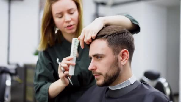 Bonito homem de olhos azuis sentado na barbearia. Cabeleireiro Cabeleireiro Mulher cortando seu cabelo. Barbeiro fêmea. — Vídeo de Stock