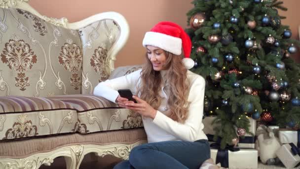 Jul kvinna klädd vit tröja Santa hatt sitter på golvet nära julgran — Stockvideo