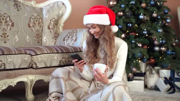 Mujer de Navidad vestido suéter blanco Santa sombrero sentado en el suelo cerca del árbol de Navidad envuelto manta sí misma — Vídeos de Stock