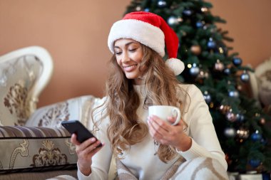 Noel kadını beyaz kazak giymiş Noel Baba şapkası takmış, Noel ağacının yanında oturuyordu. Kafkasyalı bir kadın, kış tatili ekoseli kahvesini rahatça içiyordu. Akıllı telefon kullanarak.