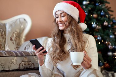 Noel kadını beyaz kazak giymiş Noel Baba şapkası takmış, Noel ağacının yanında oturuyordu. Kafkasyalı bir kadın, kış tatili ekoseli kahvesini rahatça içiyordu. Akıllı telefon kullanarak.