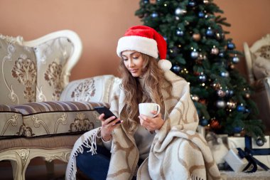 Noel kadını beyaz kazak giymiş Noel Baba şapkası takmış, Noel ağacının yanında oturuyordu. Kafkasyalı bir kadın, kış tatili ekoseli kahvesini rahatça içiyordu. Akıllı telefon kullanarak.