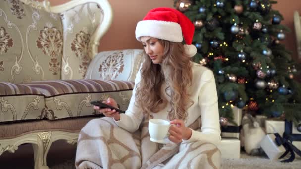 Jul kvinna klädd vit tröja Santa hatt sitter på golvet nära julgran lindade sig filt — Stockvideo