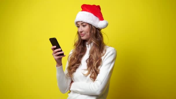Kvinna Jul Santa Hatt vit tröja gul studio bakgrund med smartphone i handen — Stockvideo