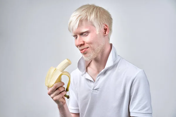 白化病白化病男子白色皮肤头发工作室穿着T恤孤立的白色背景 不正常的偏差不寻常的外表皮肤异常漂亮 外表特殊的人 吃香蕉 — 图库照片