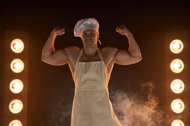 Beyaz önlük ve şef şapkası takan kaslı şefin portresi, dumanlı arka plan erkek ev kadınında güçlü pazı kaslarını gösteriyor. Kocası mutfakta. vahşi kasap..