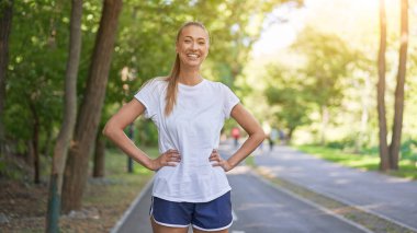 Yaz parkının önünde koşan kadın koşucu orta yaşlı atletik kadın koşuya çıkmadan önce vücut ısıtıyor koşan beyaz gömlekli şortlu koşu pistinde koşuyor.