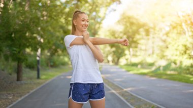 Yaz parkında spor yapmadan önce kollarını esneten kadın orta yaşlı atletik kadın koşuya çıkmadan önce beyaz şortuyla koşan beyaz şortlu koşucu ısınıyor.