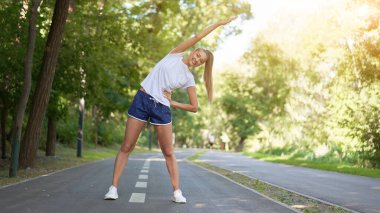 Yaz parkında spor yapmadan önce kollarını esneten kadın orta yaşlı atletik kadın koşuya çıkmadan önce beyaz şortuyla koşan beyaz şortlu koşucu ısınıyor.