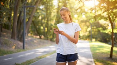 Kadın kulaklık takıp müzik dinliyor yaz parkında koşuya çıkmadan önce Kafkasyalı kadın güneşli bir sabah koşusuna hazırlanıyor koşucu elinde akıllı telefon ile antrenman yapıyor sağlıklı yaşam tarzı pozitif kavram