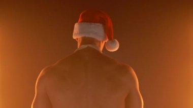Baştan çıkarıcı kaslı adam, önlüklü ve Noel Baba şapkalı ileri doğru gidiyor, kameraya dönüyor ve parmaklarıyla işaret ediyor, yavaş çekim.